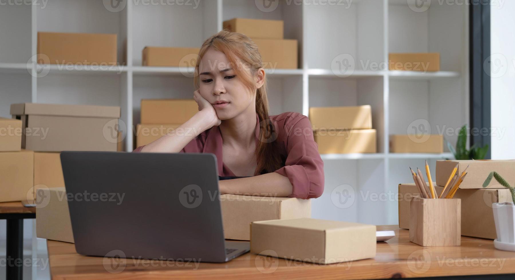 A portrait of young serious Asian woman working with laptop in the office full of packages and boxes stacking up, busy looking table, for SME, delivery, start up business and home office concept. photo