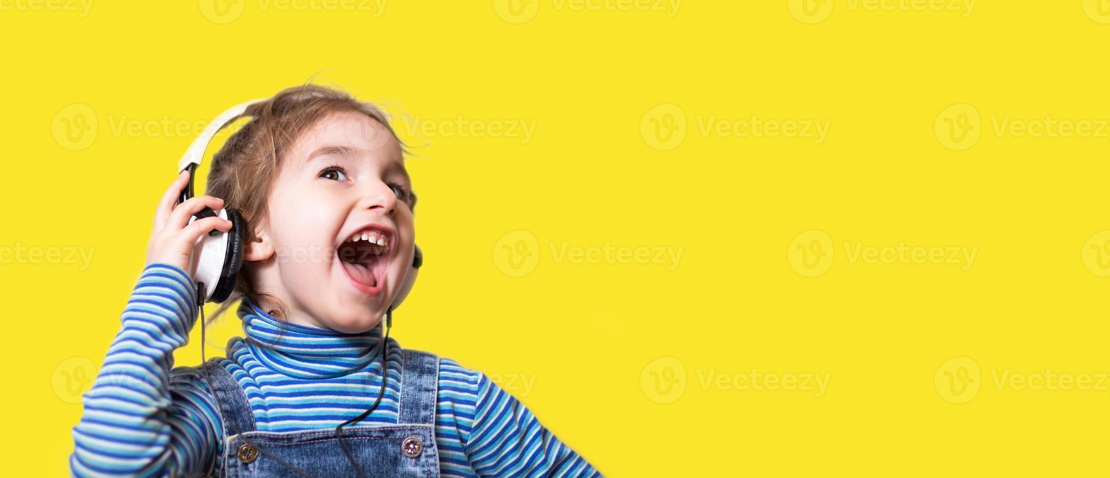 una niña con un cuello de tortuga azul a rayas escucha música con auriculares blancos y canta en voz alta con la boca abierta. diversión, gritos, emociones infantiles. fondo amarillo, pancarta, espacio de copia foto