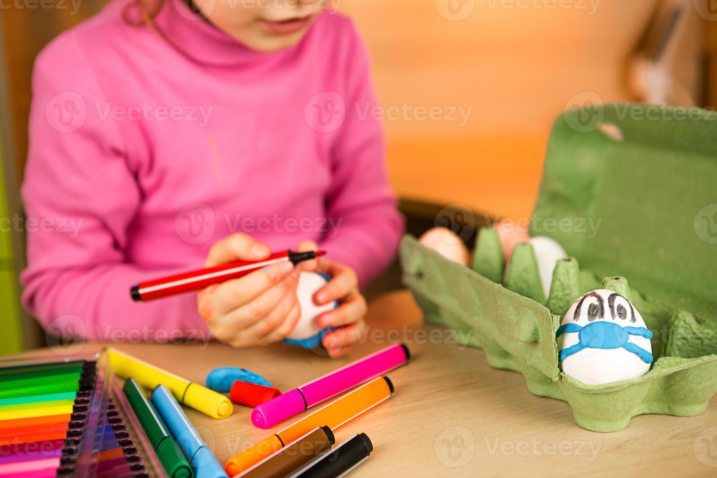 Girl with bunny ears makes medical masks out of plasticine for Easter  eggs. An egg with eyes and a blue mask in the tray. DIY sitting at home, preparing for a religious holiday, home interior. photo