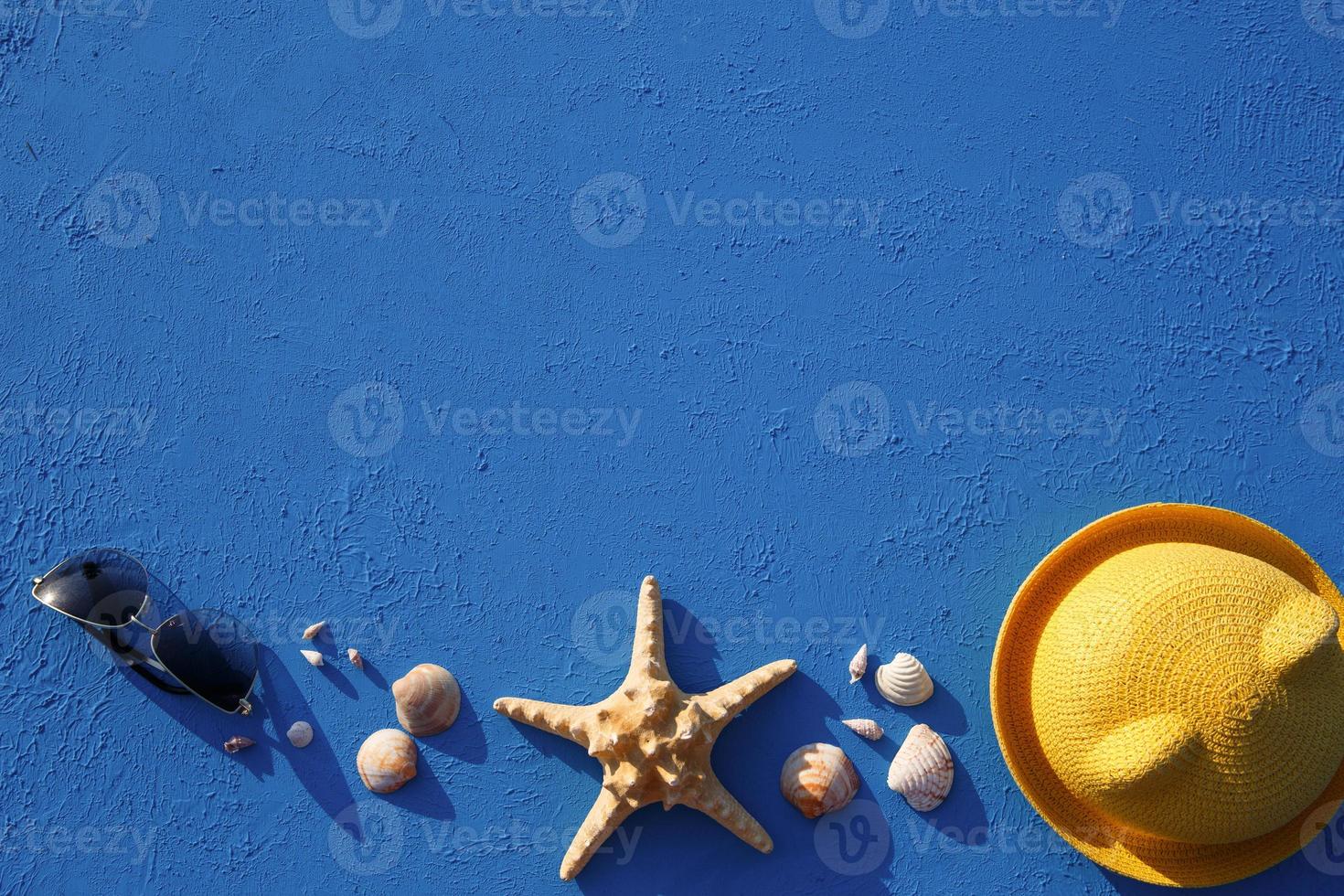 Frame with beach accessories on a nautical theme yellow straw hat, sunglasses, starfish and shells on a blue background. Vacation concept, sea trip, UV protection, swimming. Copy space. Flatlay photo