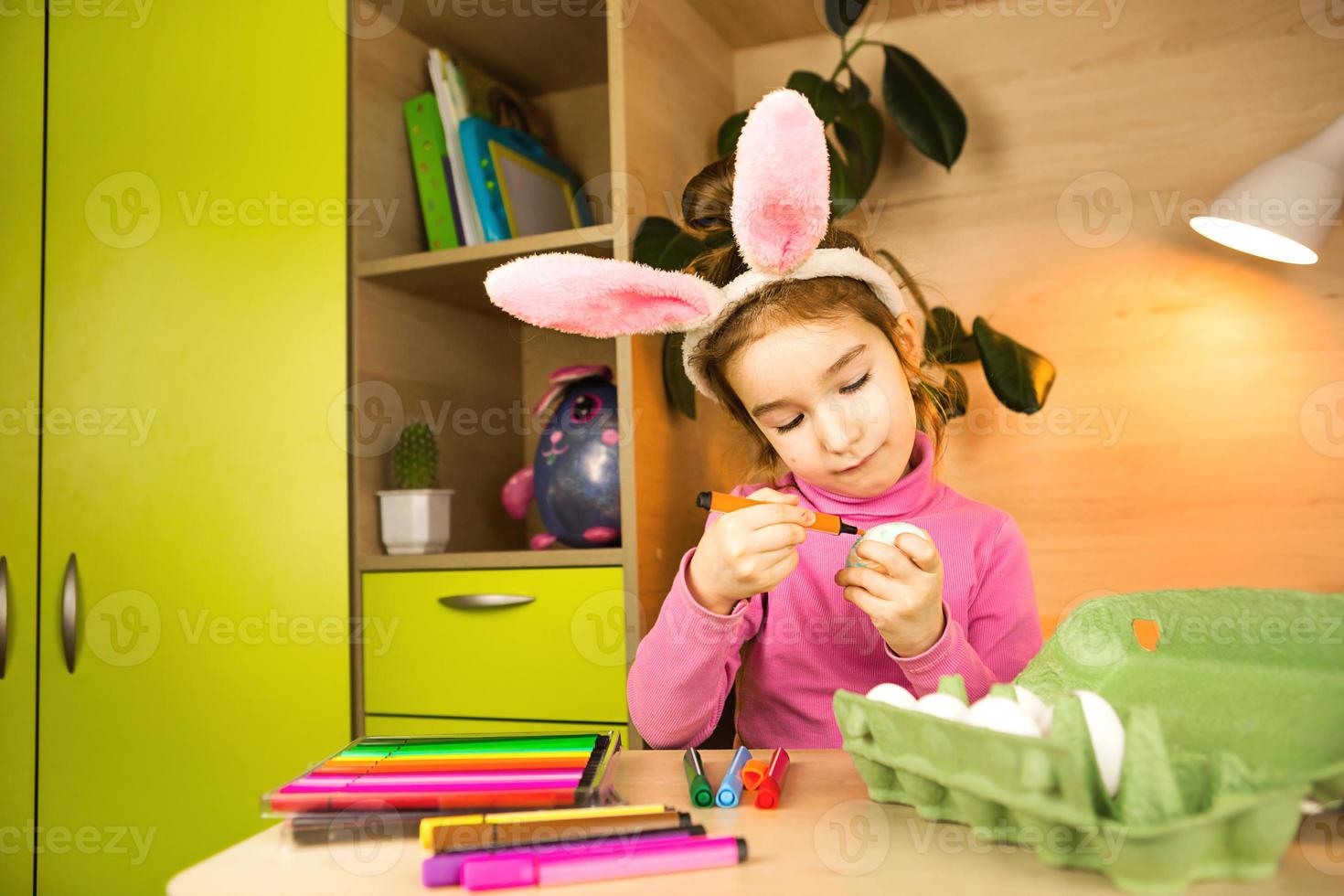 A girl in the ears of an Easter bunny paints eggs with a felt-tip pen in the home interior. Crafts, preparation for a religious holiday, a tray with eggs, hare ears made of plasticine photo