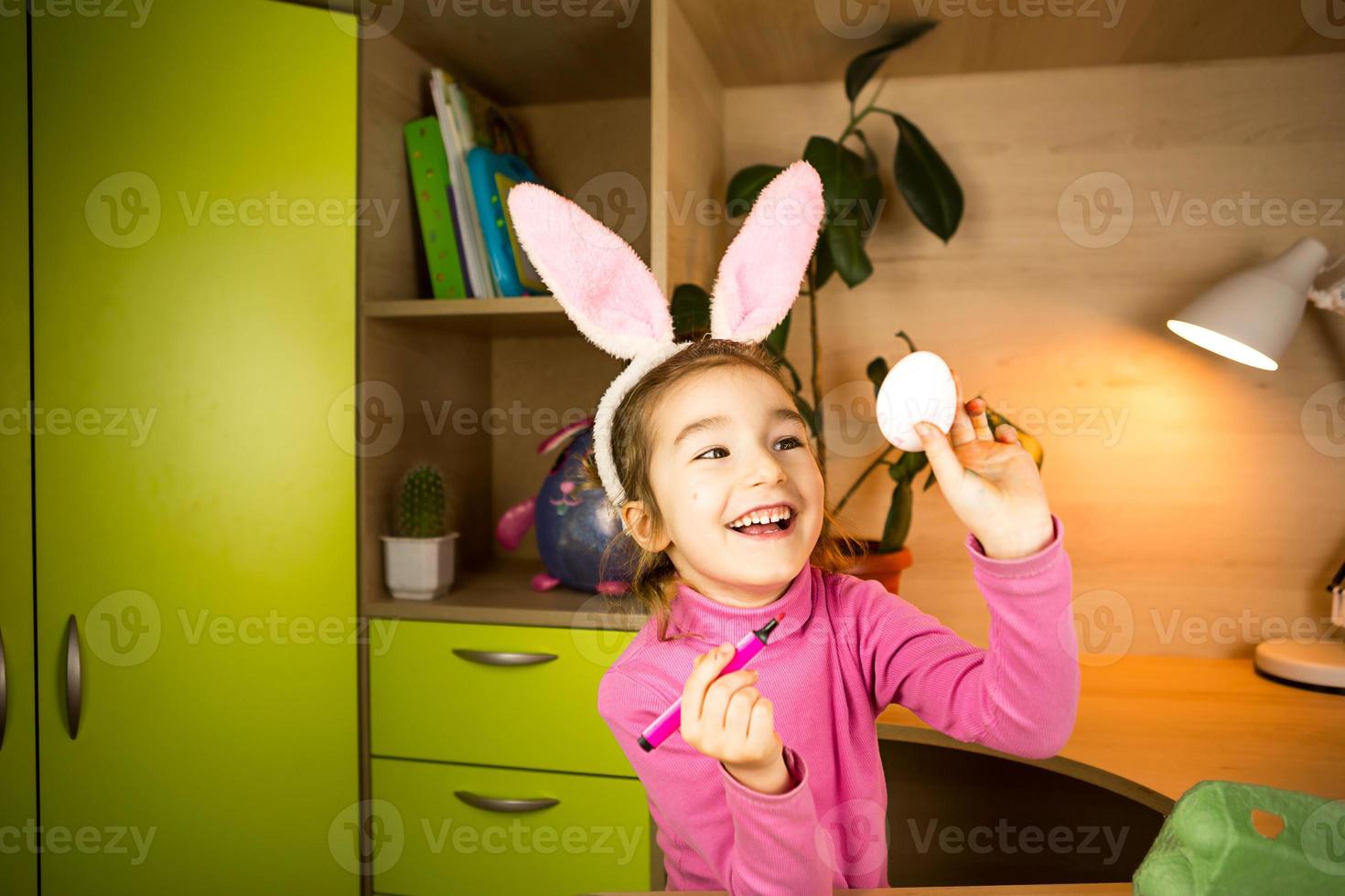 A girl in the ears of an Easter bunny paints eggs with a felt-tip pen in the home interior. Crafts, preparation for a religious holiday, a tray with eggs, hare ears made of plasticine photo
