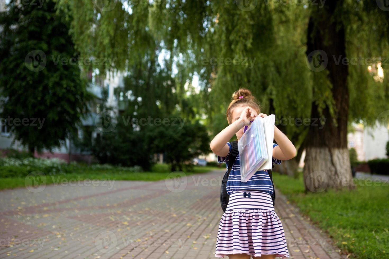 A little girl of Caucasian appearance in a school uniform with a backpack and the book and a set of markers. back to school. Elementary school, developing activities for preschoolers. Space for text photo