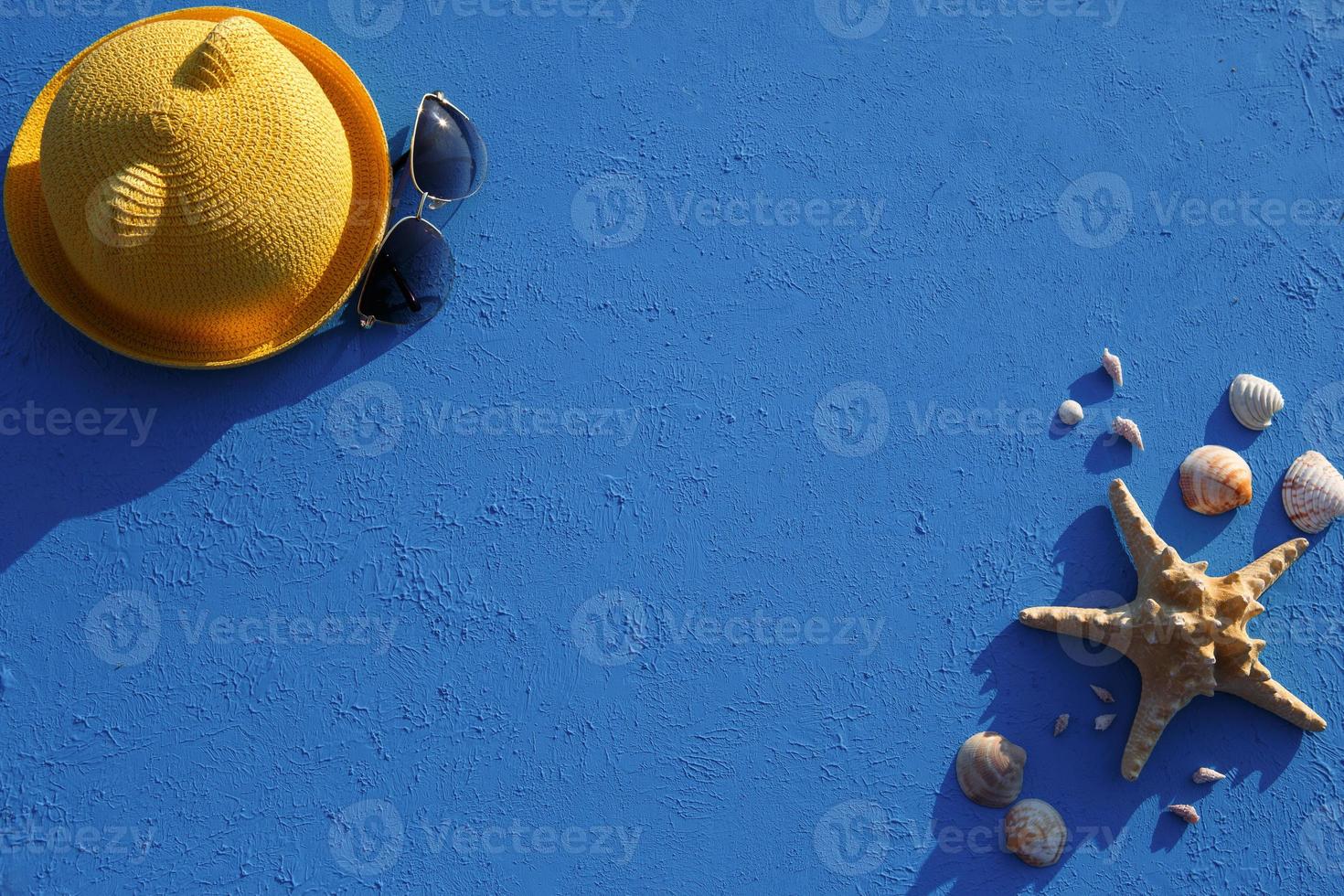 Frame with beach accessories on a nautical theme yellow straw hat, sunglasses, starfish and shells on a blue background. Vacation concept, sea trip, UV protection, swimming. Copy space. Flatlay photo