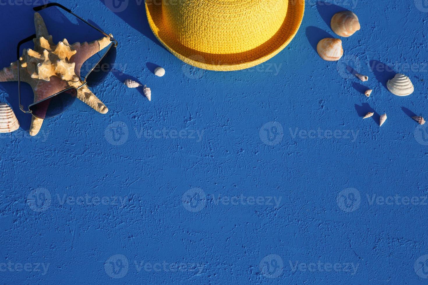 Frame with beach accessories on a nautical theme yellow straw hat, sunglasses, starfish and shells on a blue background. Vacation concept, sea trip, UV protection, swimming. Copy space. Flatlay photo