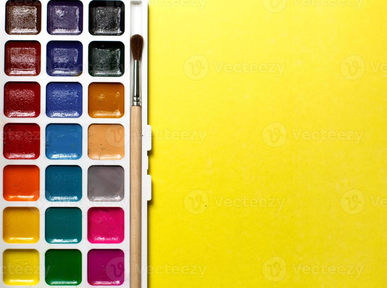 A palette of eighteen-color watercolors and paintbrushes on a yellow background with space for text. photo