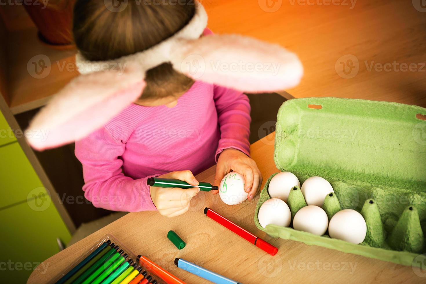 A girl in the ears of an Easter bunny paints eggs with a felt-tip pen in the home interior. Crafts, preparation for a religious holiday, a tray with eggs, hare ears made of plasticine photo