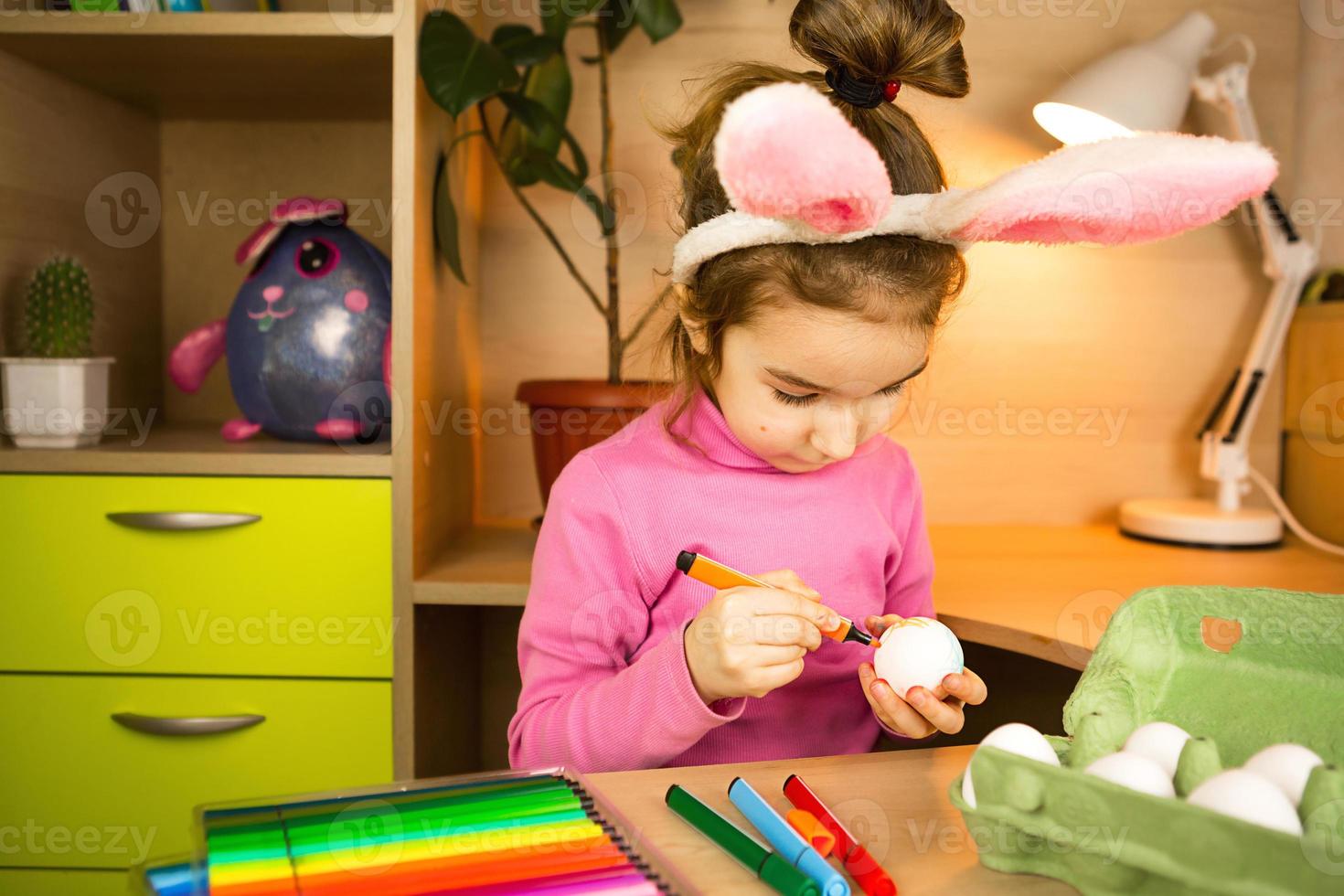 A girl in the ears of an Easter bunny paints eggs with a felt-tip pen in the home interior. Crafts, preparation for a religious holiday, a tray with eggs, hare ears made of plasticine photo