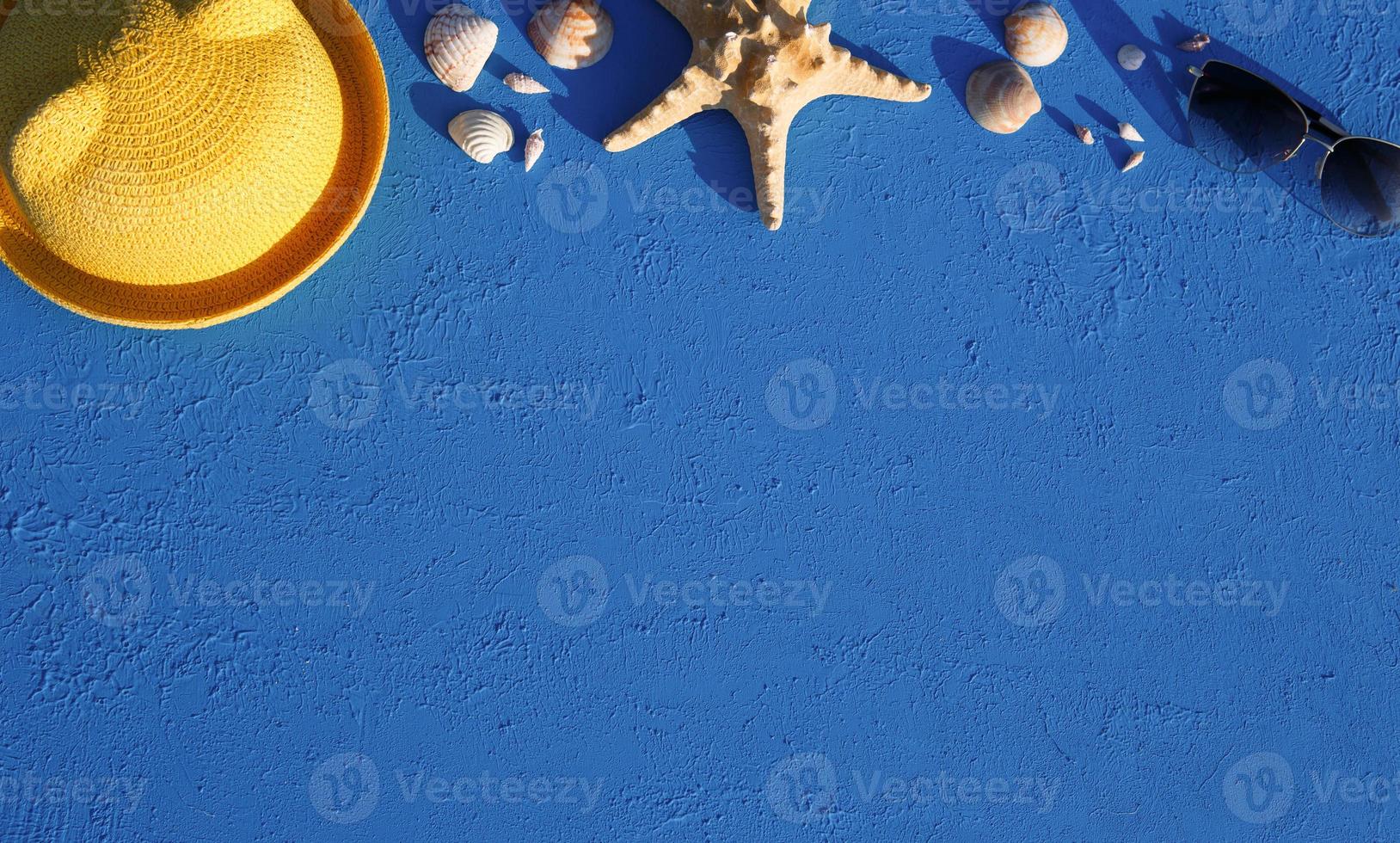 Frame with beach accessories on a nautical theme yellow straw hat, sunglasses, starfish and shells on a blue background. Vacation concept, sea trip, UV protection, swimming. Copy space. Flatlay photo