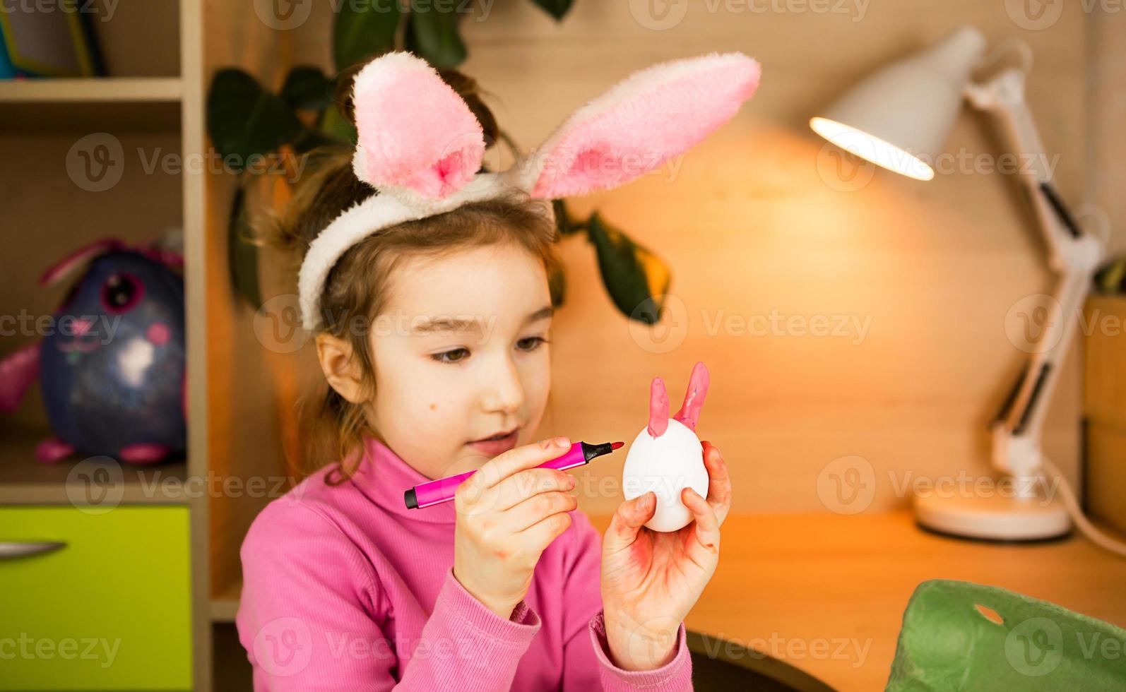 A girl in the ears of an Easter bunny paints eggs with a felt-tip pen in the home interior. Crafts, preparation for a religious holiday, a tray with eggs, hare ears made of plasticine photo