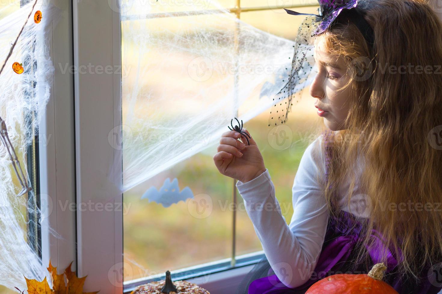 A girl in a witch costume on the windowsill by the window is playing with a spider web and a spider, decorating the house for the Halloween holiday. Terrible scenery, fear and horror, pumpkins photo