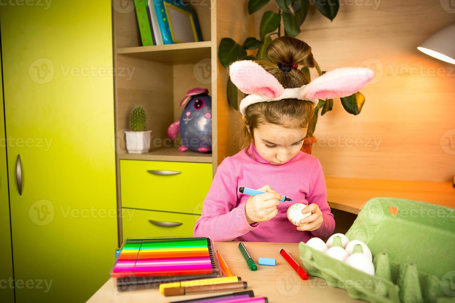 una niña en los oídos de un conejito de pascua pinta huevos con un rotulador en el interior de la casa. manualidades, preparación para una fiesta religiosa, una bandeja con huevos, orejas de liebre hechas de plastilina foto