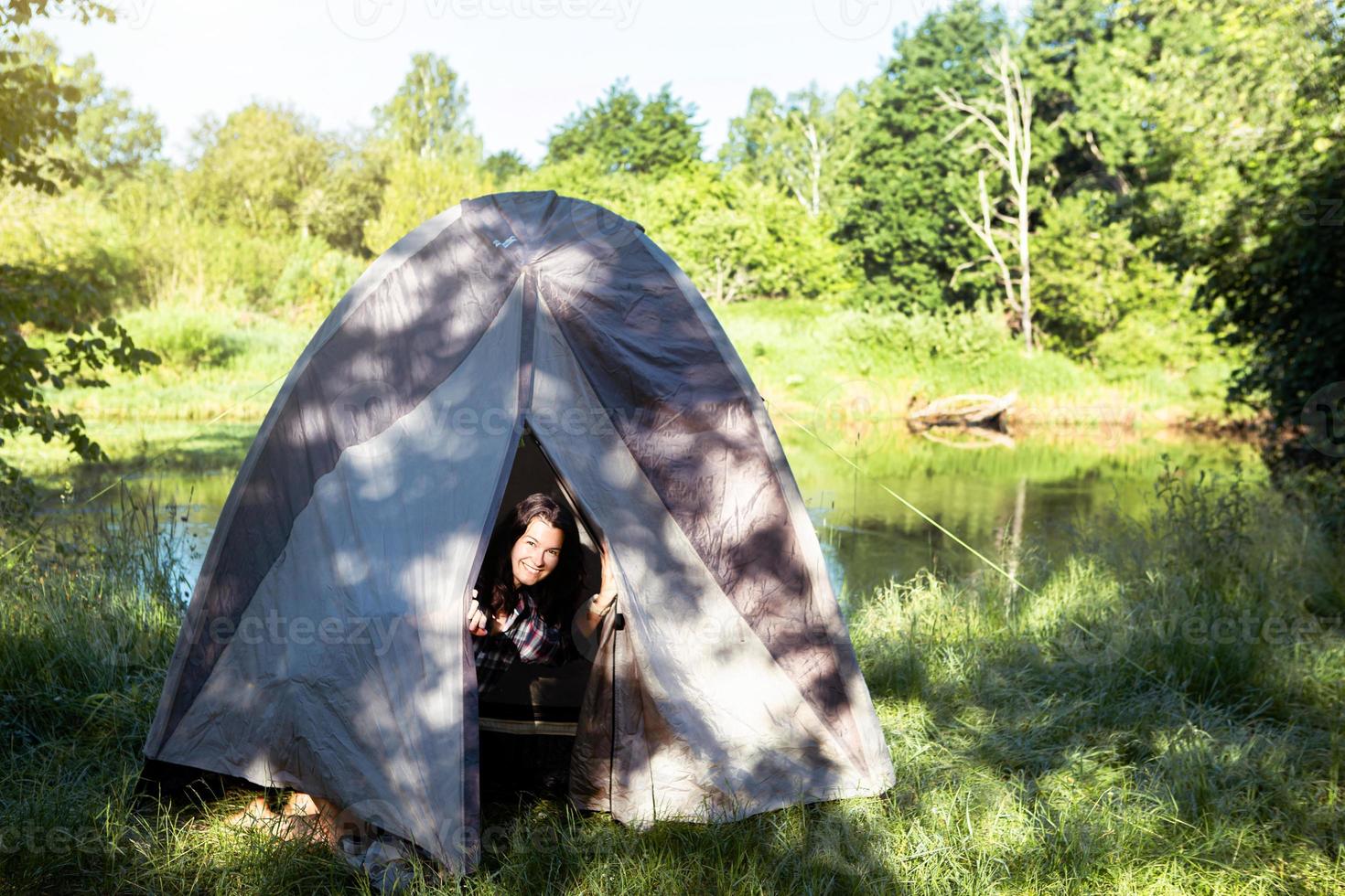 A happy woman in a plaid shirt looks out of a tourist tent on a hike on the riverbank in the morning. Camping in nature, overnight in the wild, family holidays and adventures. photo