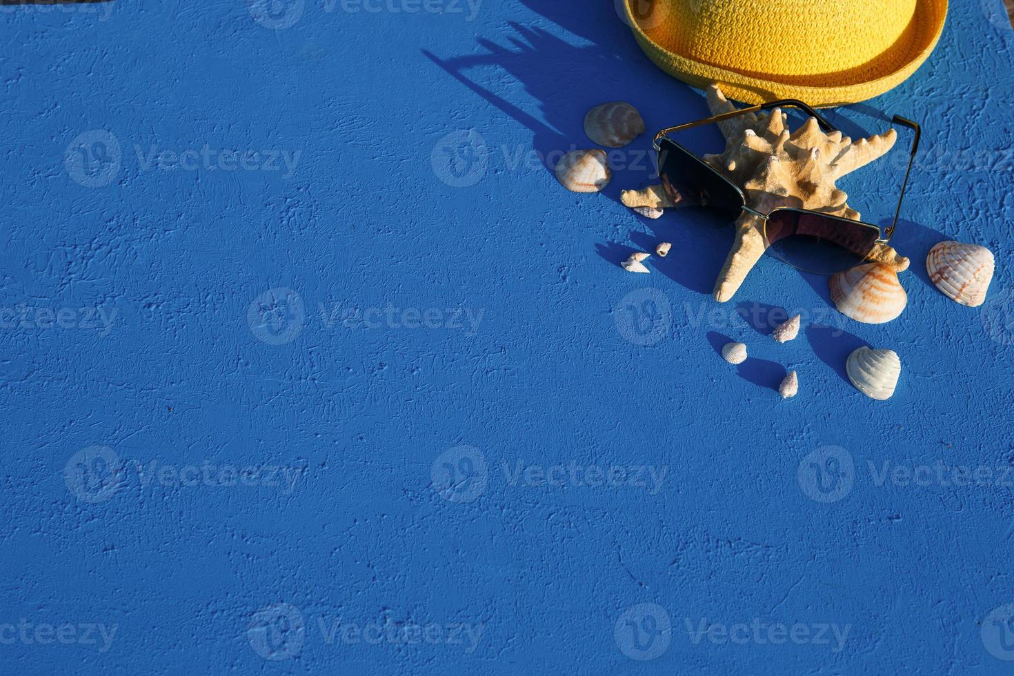 Frame with beach accessories on a nautical theme yellow straw hat, sunglasses, starfish and shells on a blue background. Vacation concept, sea trip, UV protection, swimming. Copy space. Flatlay photo