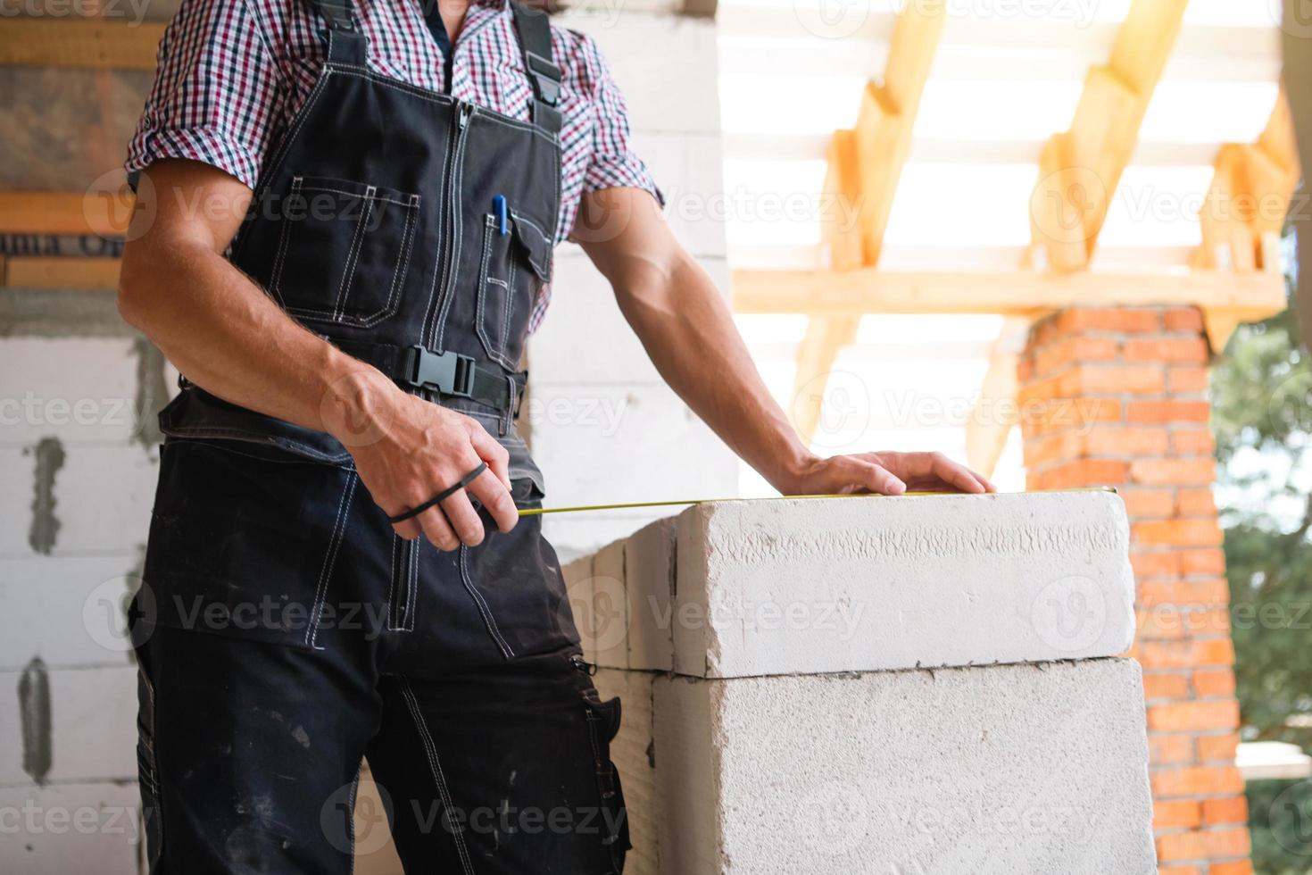 Construction worker at construction site measures the length of window opening and brick wall with tape measure. Cottage are made of porous concrete blocks, work clothes - jumpsuit and baseball cap photo