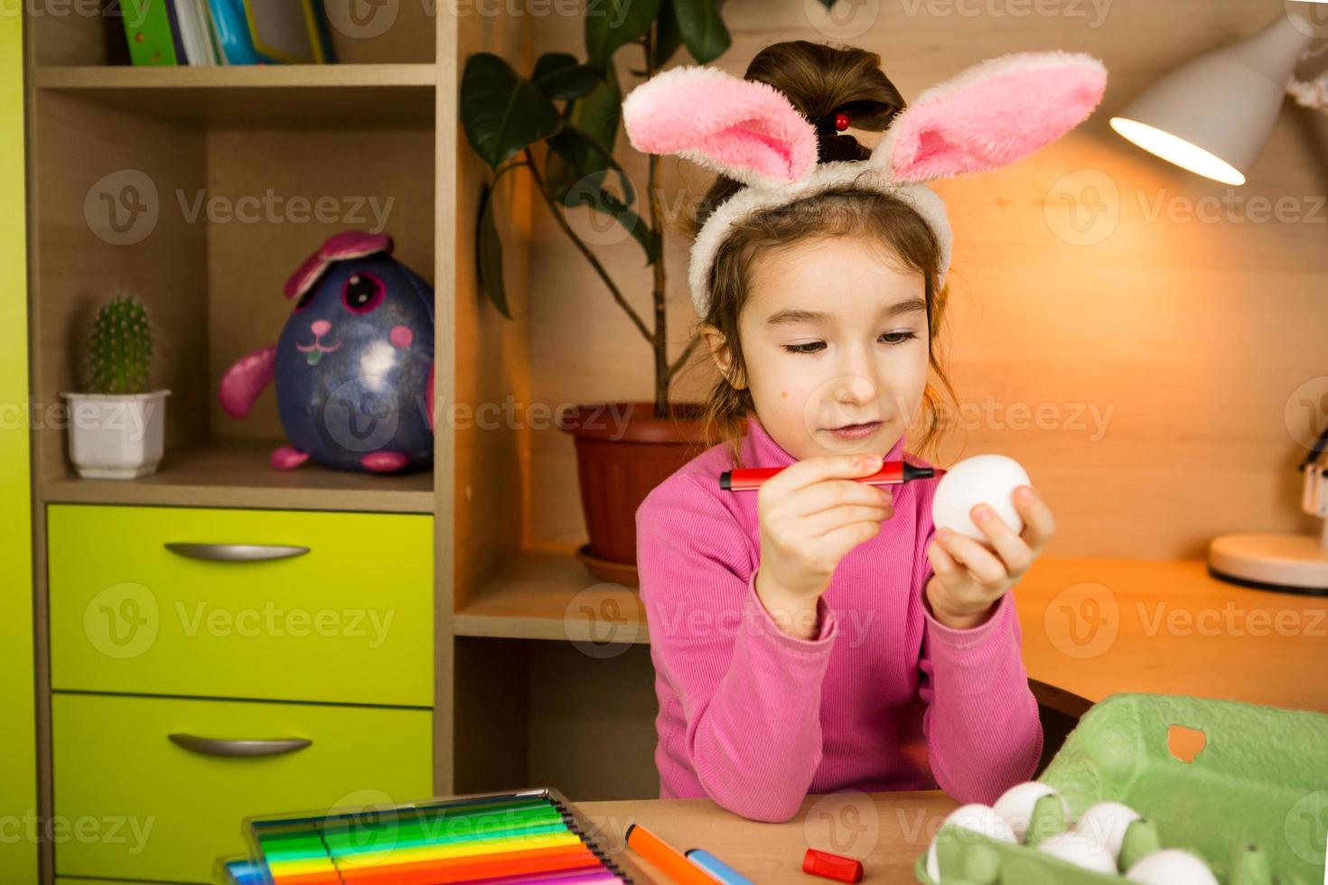 A girl in the ears of an Easter bunny paints eggs with a felt-tip pen in the home interior. Crafts, preparation for a religious holiday, a tray with eggs, hare ears made of plasticine photo