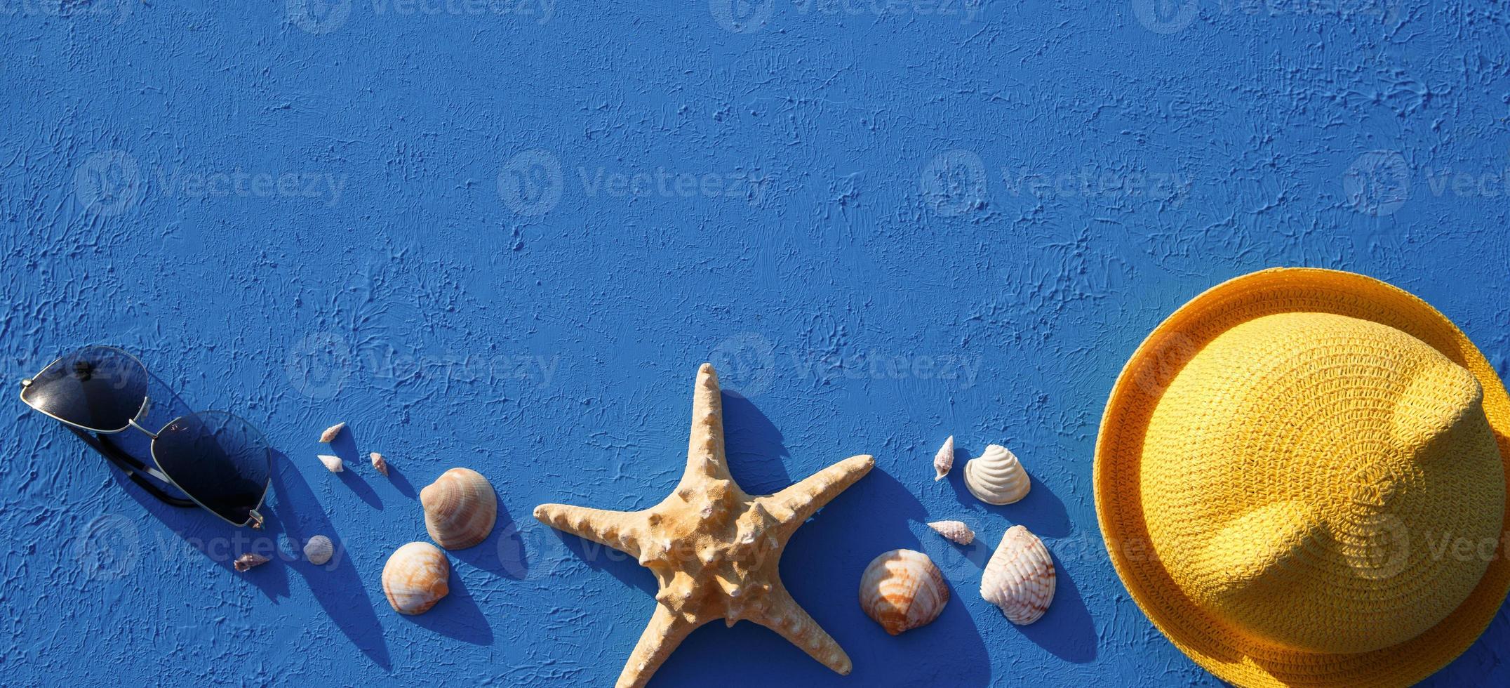 Frame with beach accessories on a nautical theme yellow straw hat, sunglasses, starfish and shells on a blue background. Vacation concept, sea trip, UV protection, swimming. Copy space. Flatlay photo