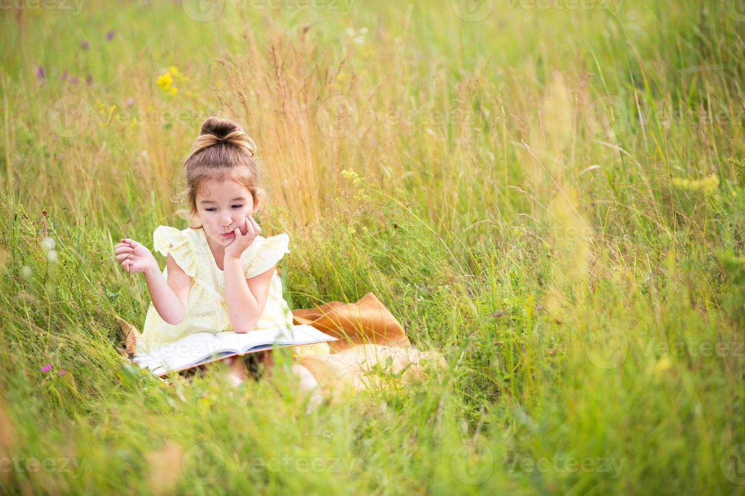 Girl in a yellow dress sits in the grass on a blanket in a field and reads a paper book. International Children's Day. Summer time, childhood, education and entertainment, cottage core. Copy space photo