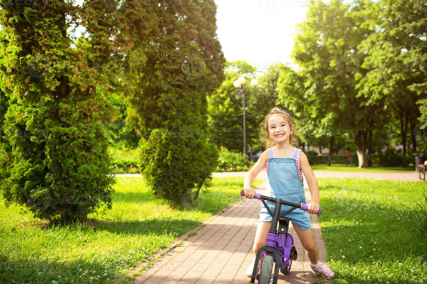 A girl in a denim jumpsuit rides a purple running bike in the summer in a green park. Children's active entertainment, scooter for toddlers, happy baby. Copy space photo