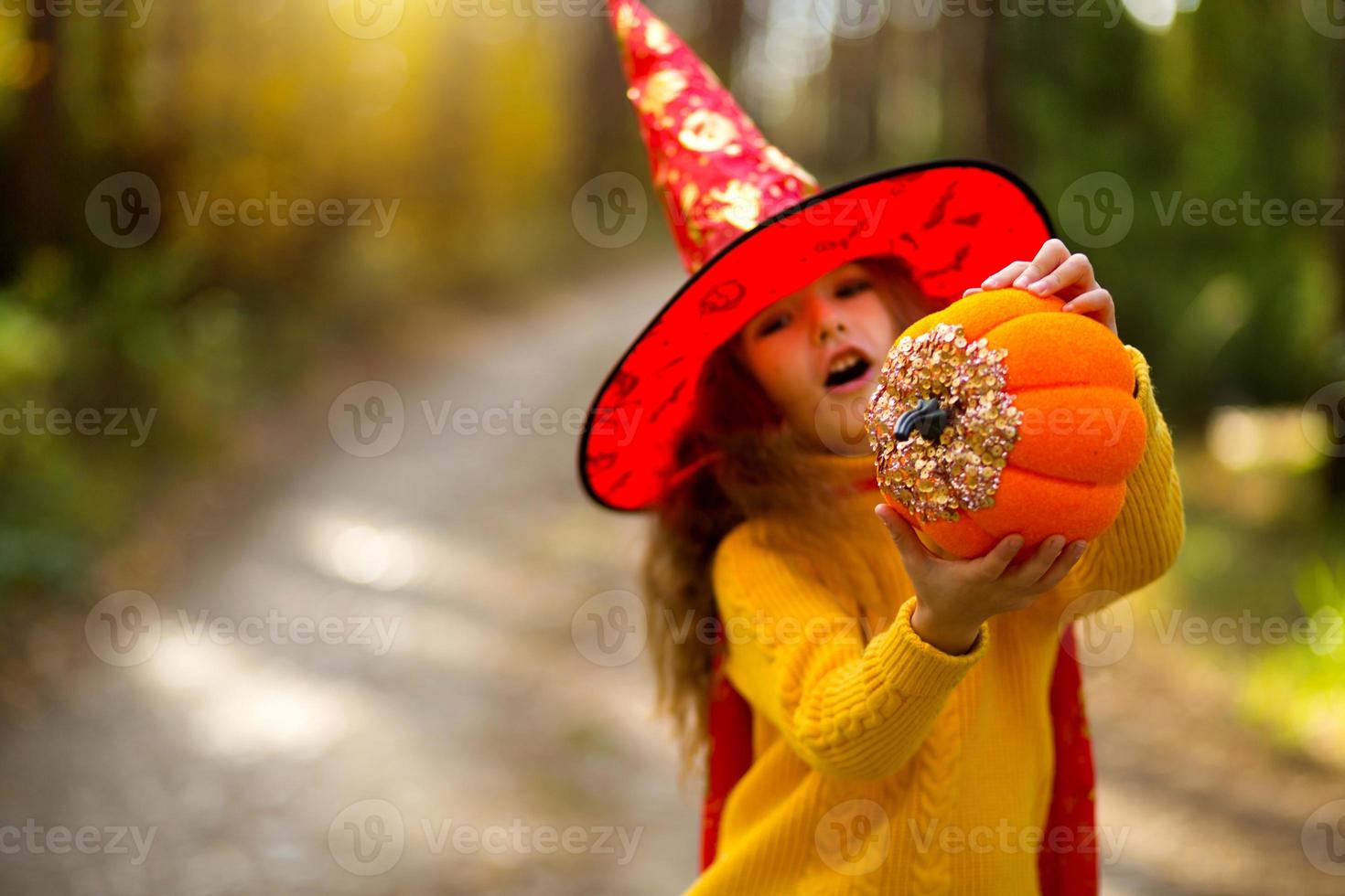 A girl in a witch's hat, in a yellow sweater and with a pumpkin with carved eyes and a mouth Jack o Lantern in a yellow autumn forest. Halloween Holiday photo