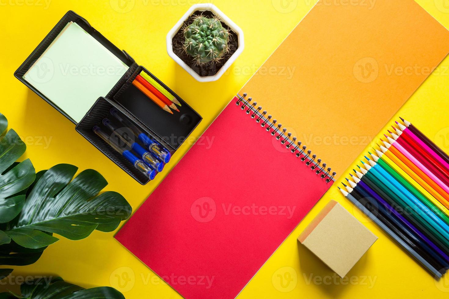Layout of multi-colored stationery on a yellow background spiral notebook, colored pencils, stand, pens, indoor flower. Business flat lay, mock up. Sheets for notes, office work. Back to school photo
