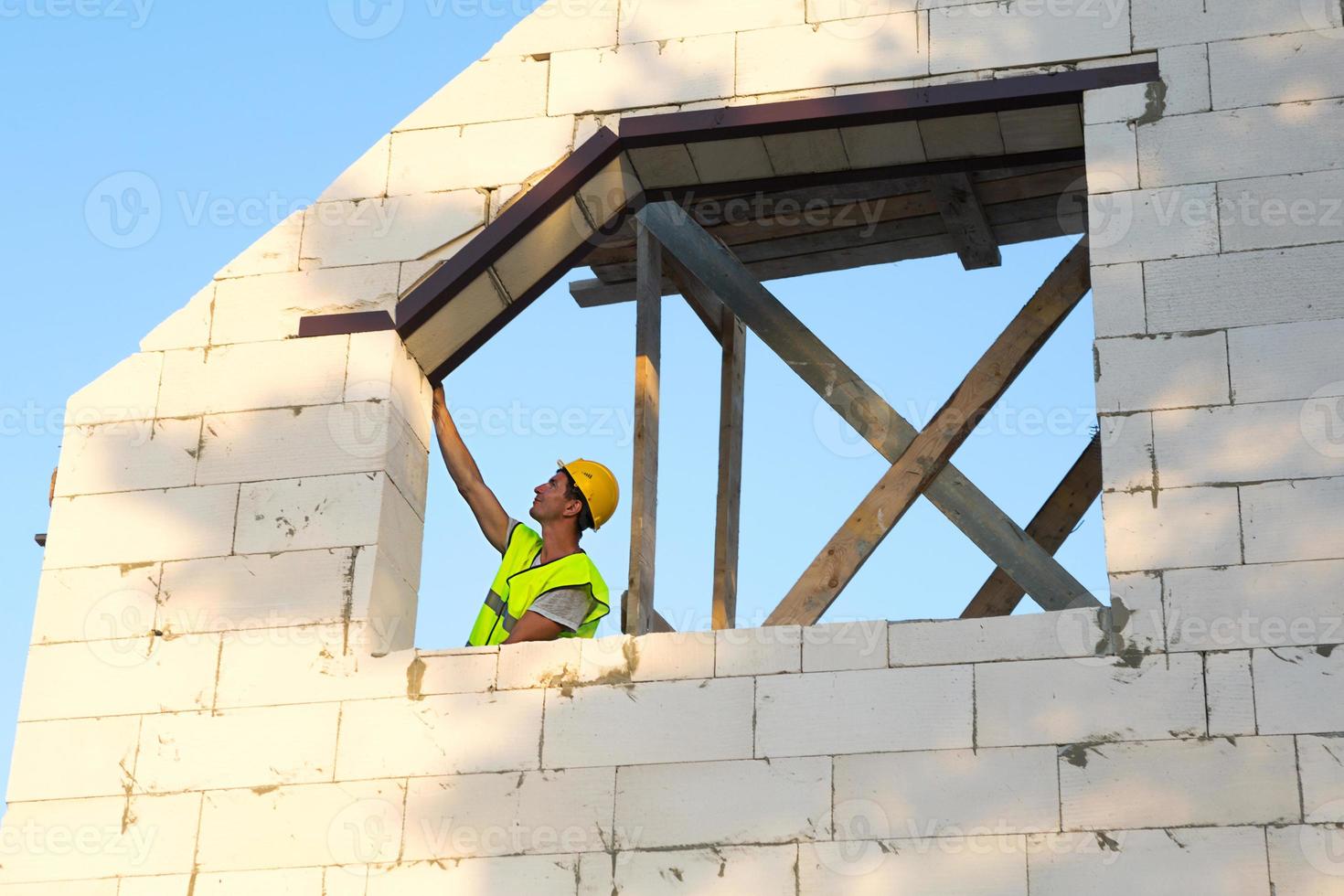 trabajador de la construcción en el sitio de construcción mide la longitud de la abertura de la ventana y la pared con cinta métrica. la cabaña está hecha de bloques de hormigón poroso, ropa protectora: casco y chaleco foto