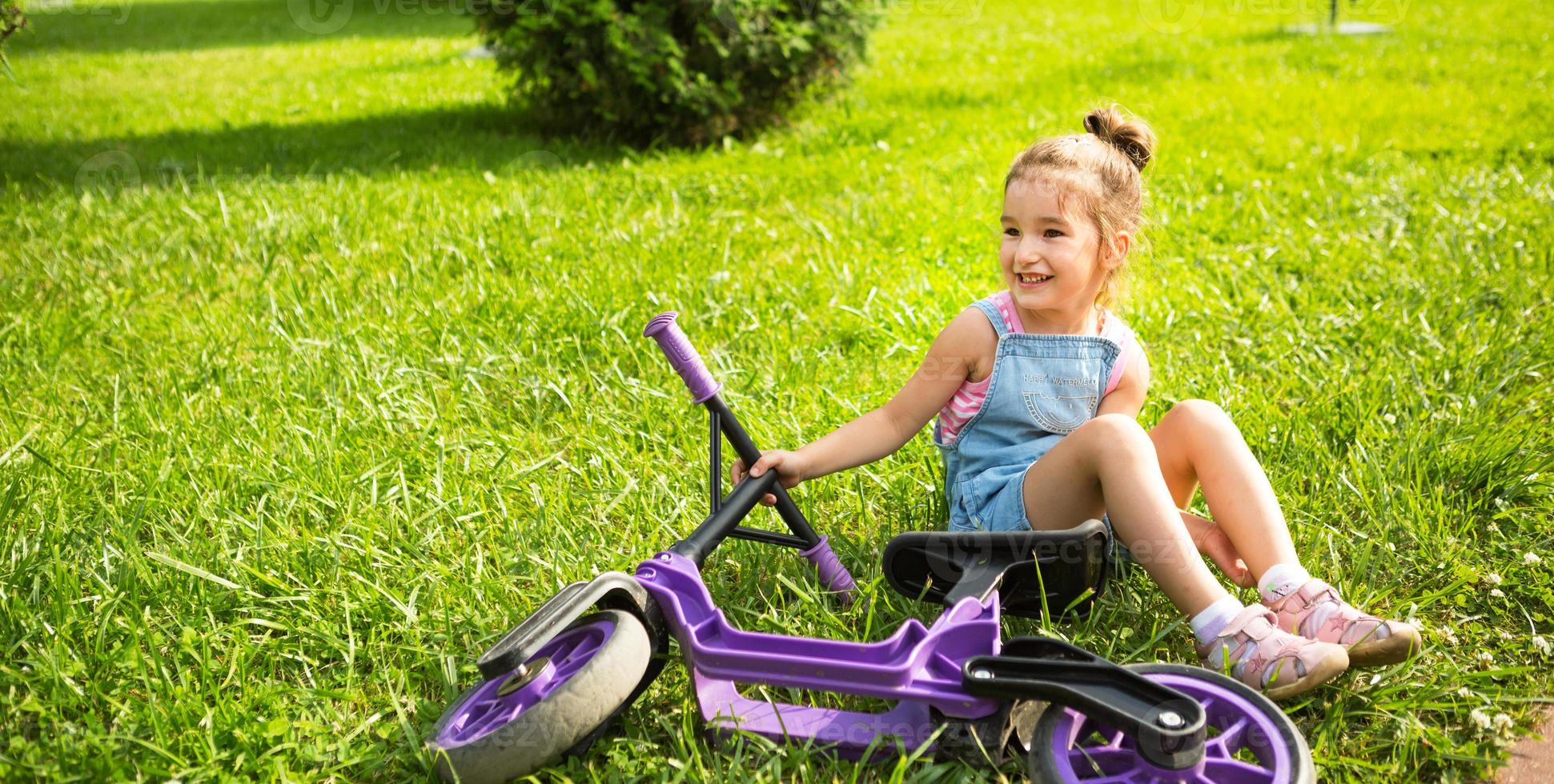 Cheerful girl sitting resting on the grass in a park near to the purple running bike in summer time. Children's active entertainment, scooter for toddlers, happy child. Copy space. Summer background photo