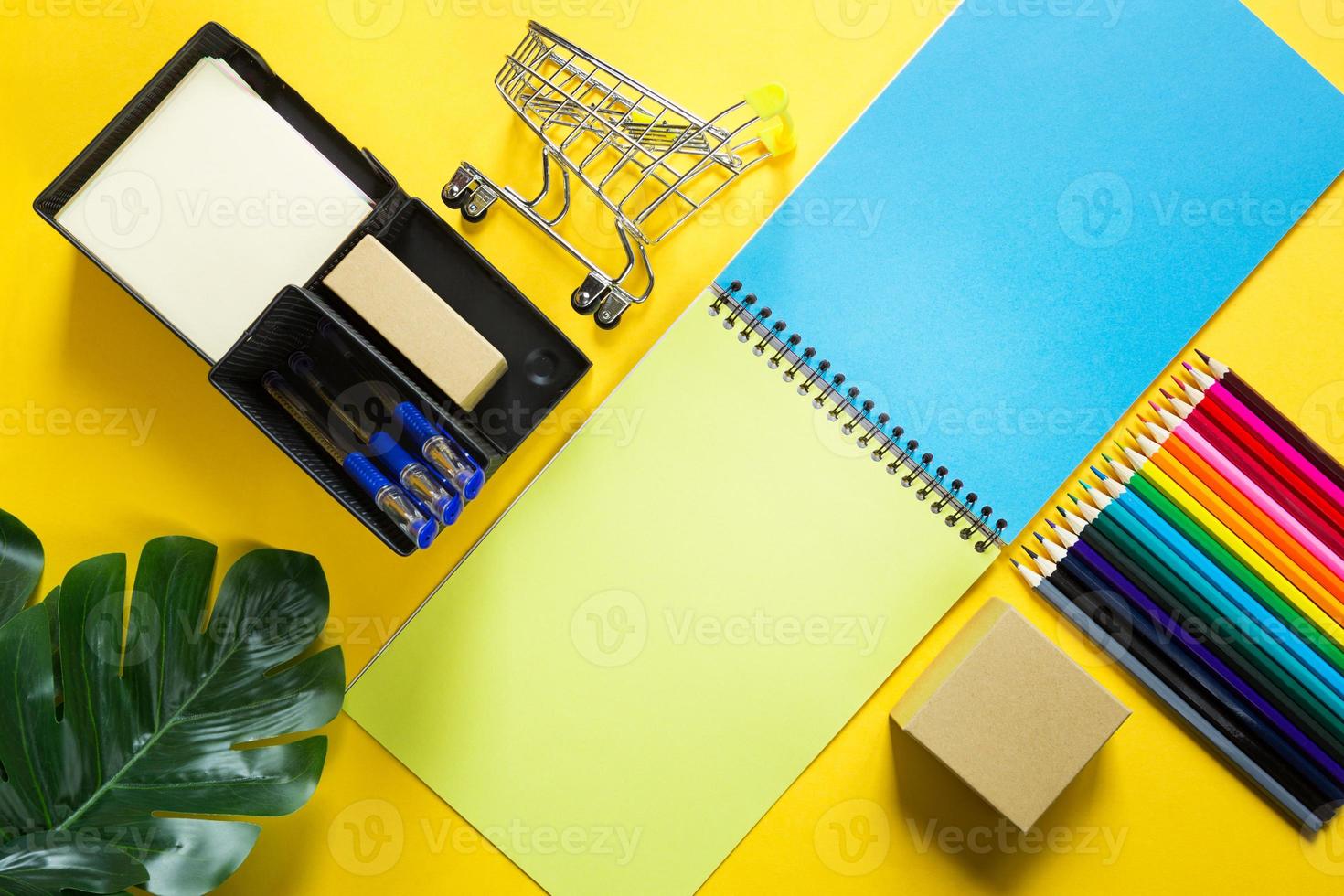 Layout of multi-colored stationery on a yellow background spiral notebook, colored pencils, stand, pens, indoor flower. Business flat lay, mock up. Sheets for notes, office work. Back to school photo