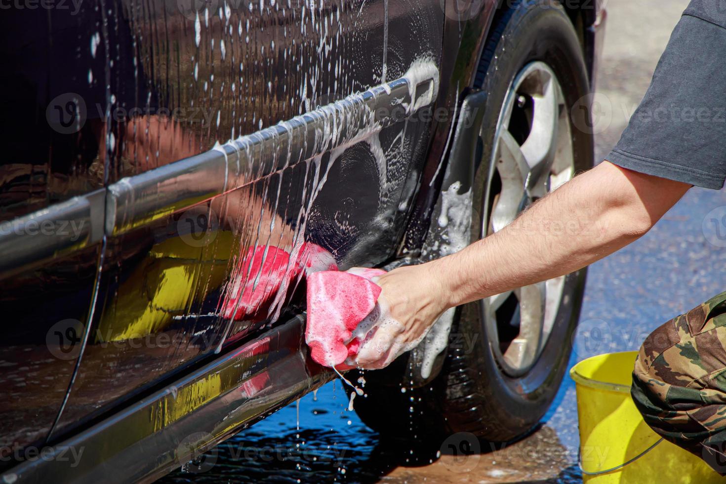 el hombre lava la rueda del coche. la mano masculina sostiene una esponja rosa con espuma jabonosa para limpiar. foto