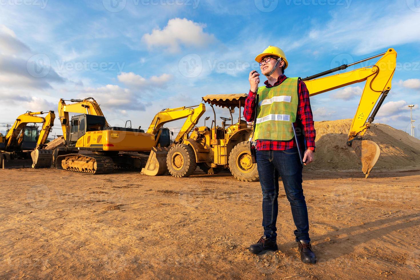 asian technician civil engineer talk with team by walkie talkie hold laptop computer or tablet for transport construction work project with excavator on engineering site background photo