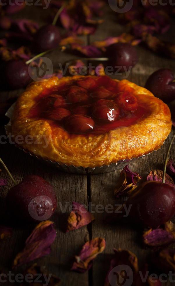 Tarta de mermelada de cereza con cerezas crudas colocadas alrededor y rosas secas sobre tablas de madera sobre fondo negro. foto