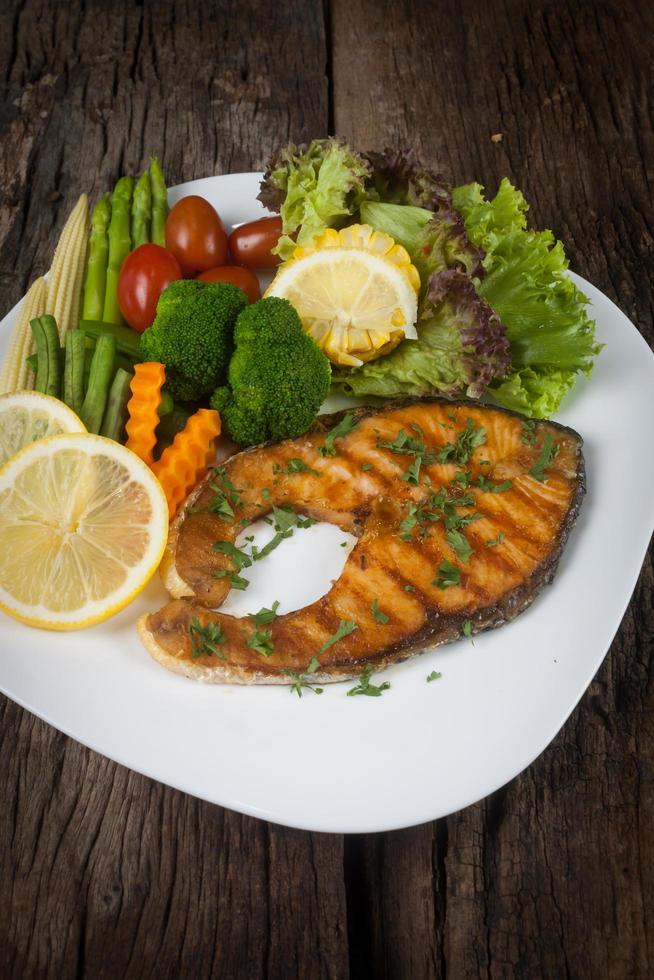 Salmon steak with sauce on a white plate with lemon on plate and crispy French fries. Many vegetables are placed around the dish on the wooden floor. photo