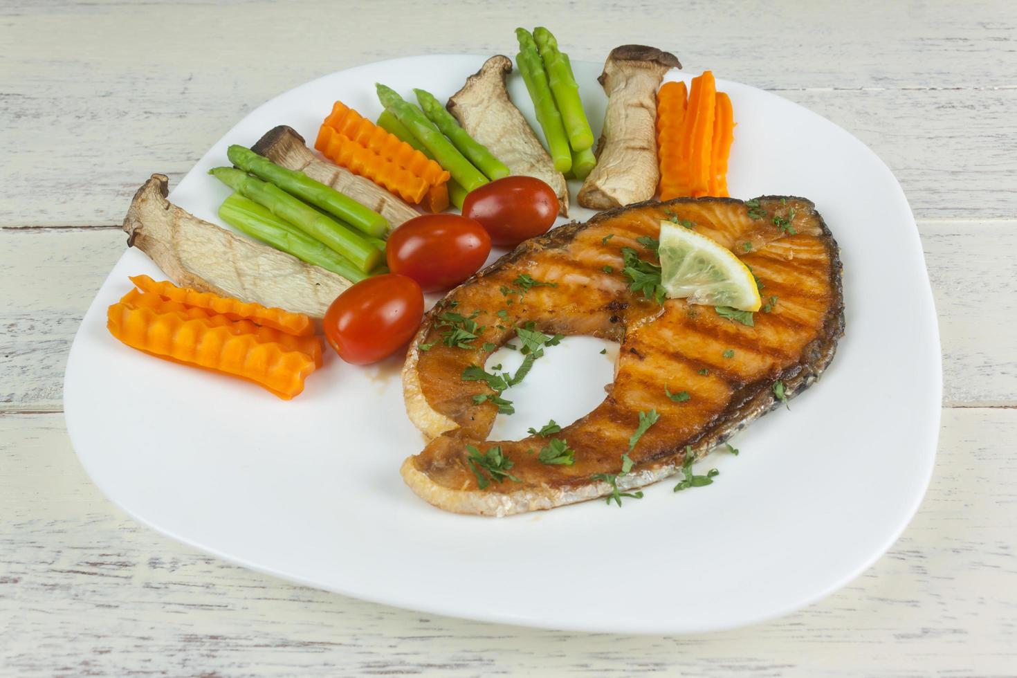 Grilled salmon steak slices until cooked, placed on a white plate with lemon on fish and vegetables, placed around the plate on a white wooden floor. photo