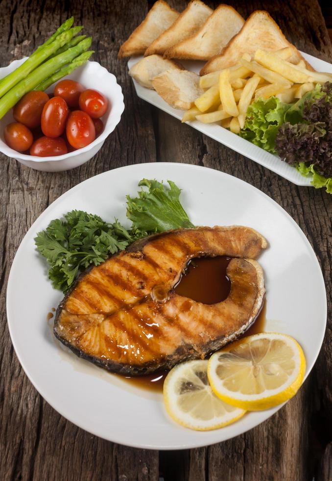 bistec de salmón con salsa en un plato blanco con limón en el plato y papas fritas crujientes. muchas verduras se colocan alrededor del plato en el suelo de madera. foto