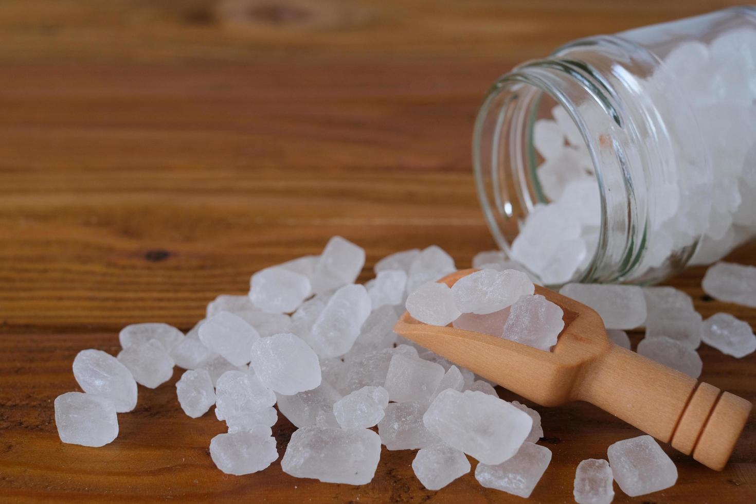 White candy rock sugar or or crystal sugar in wooden scoop and glass jar isolated on wooden background. White rock sugar is melted and then allowed to crystallize into semi-translucent gemstone. photo