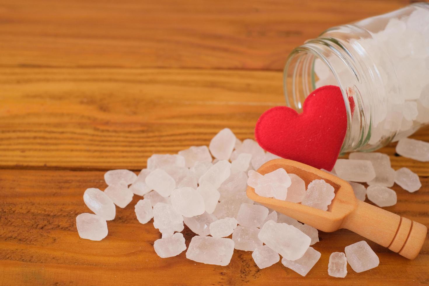 White candy rock sugar, crystal sugar in wooden scoop and glass jar with red hearts on wooden background. Selected focus. photo