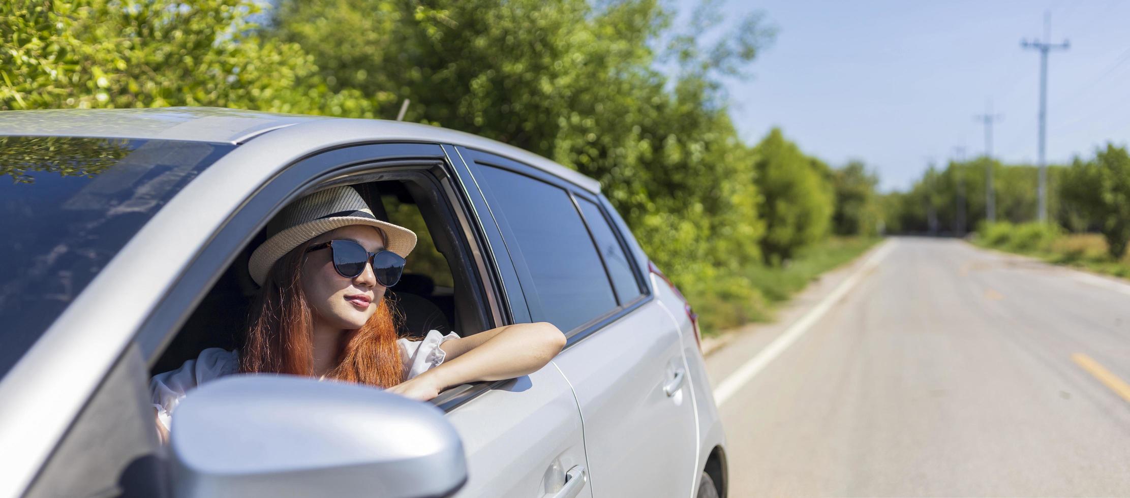 Woman with ginger red hair wearing sunglasses driving car in solo weekend getaway vacation trip looking out the window in summer with lush green nature in countryside road for freedom and adventure photo