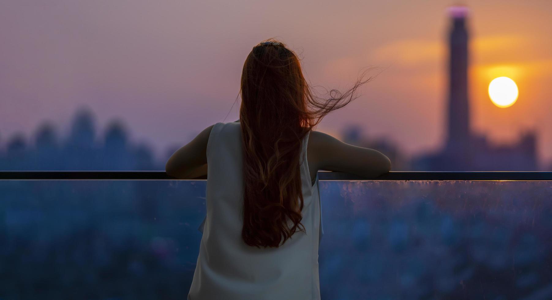 Woman looking and enjoying the sunset view from balcony with the sun setting behind skyscraper in busy urban downtown with loneliness for solitude, loneliness and dreaming of freedom lifestyle photo