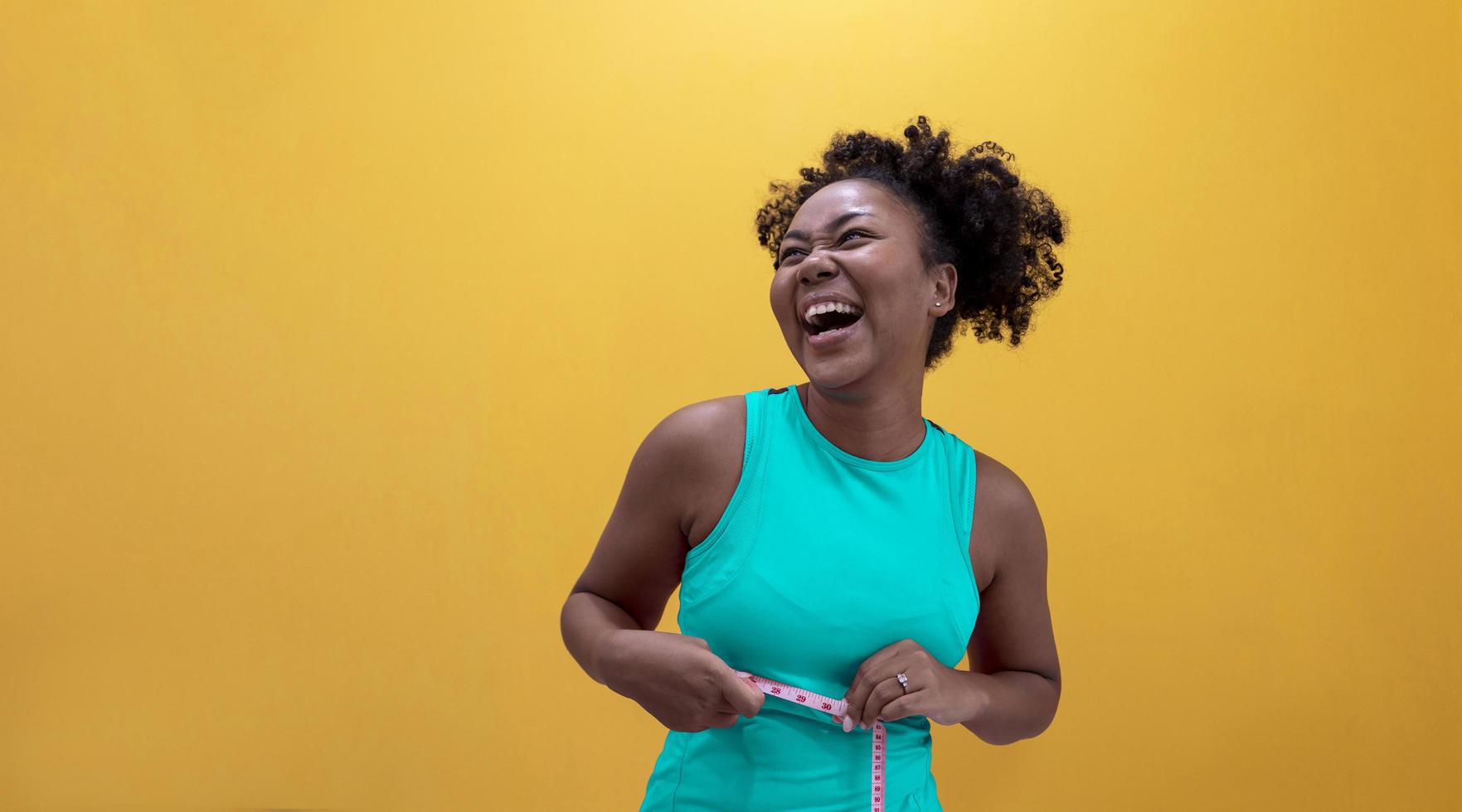 Happy African American woman in sportswear is happy while measuring waist with tape isolated on yellow background for healthy food and exercise concept photo