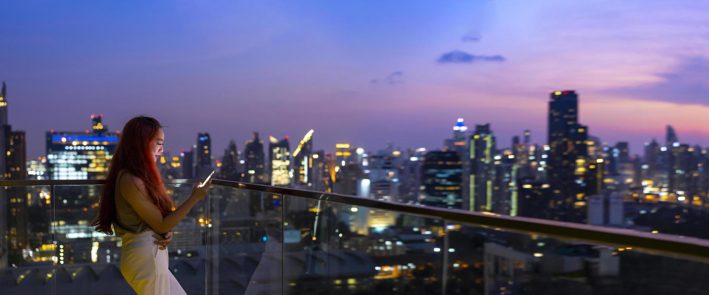 Panorama of Asian woman using mobile phone with Bangkok skyscraper cityscape at sunset blue hour at downtown urban district for modern luxury living and prestige travel concept photo