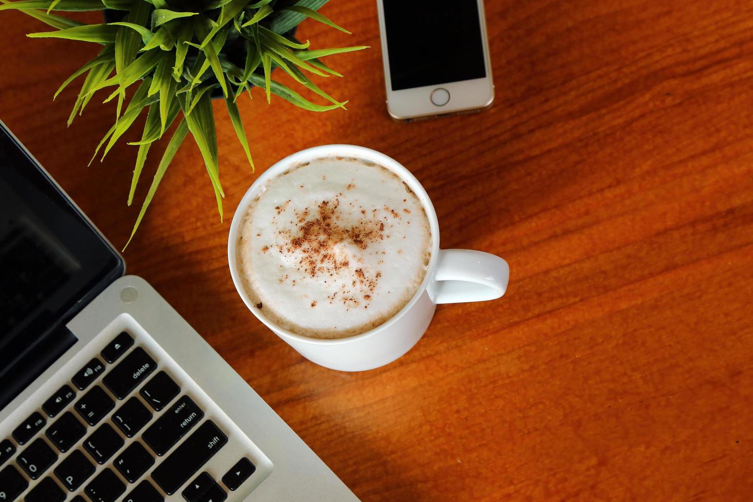 computadora portátil, teléfono inteligente y una taza de café con leche caliente en la vista superior de la mesa de madera. foto