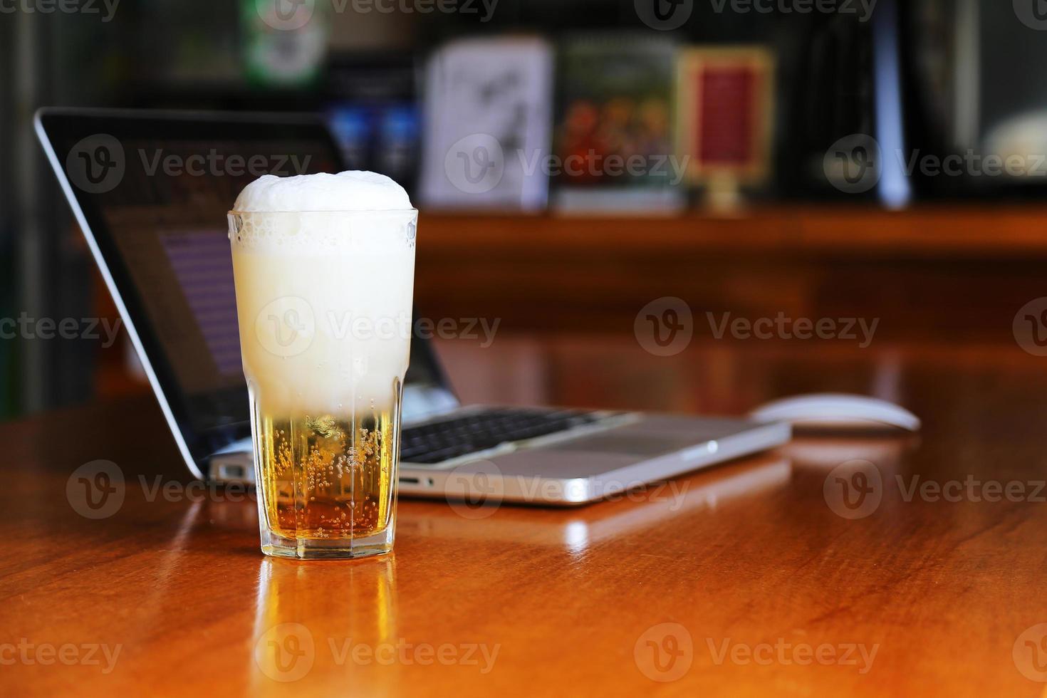 Glass of beer on wooden table with computer notebook in cafe. Beer in mug on table with laptop background in bar. photo