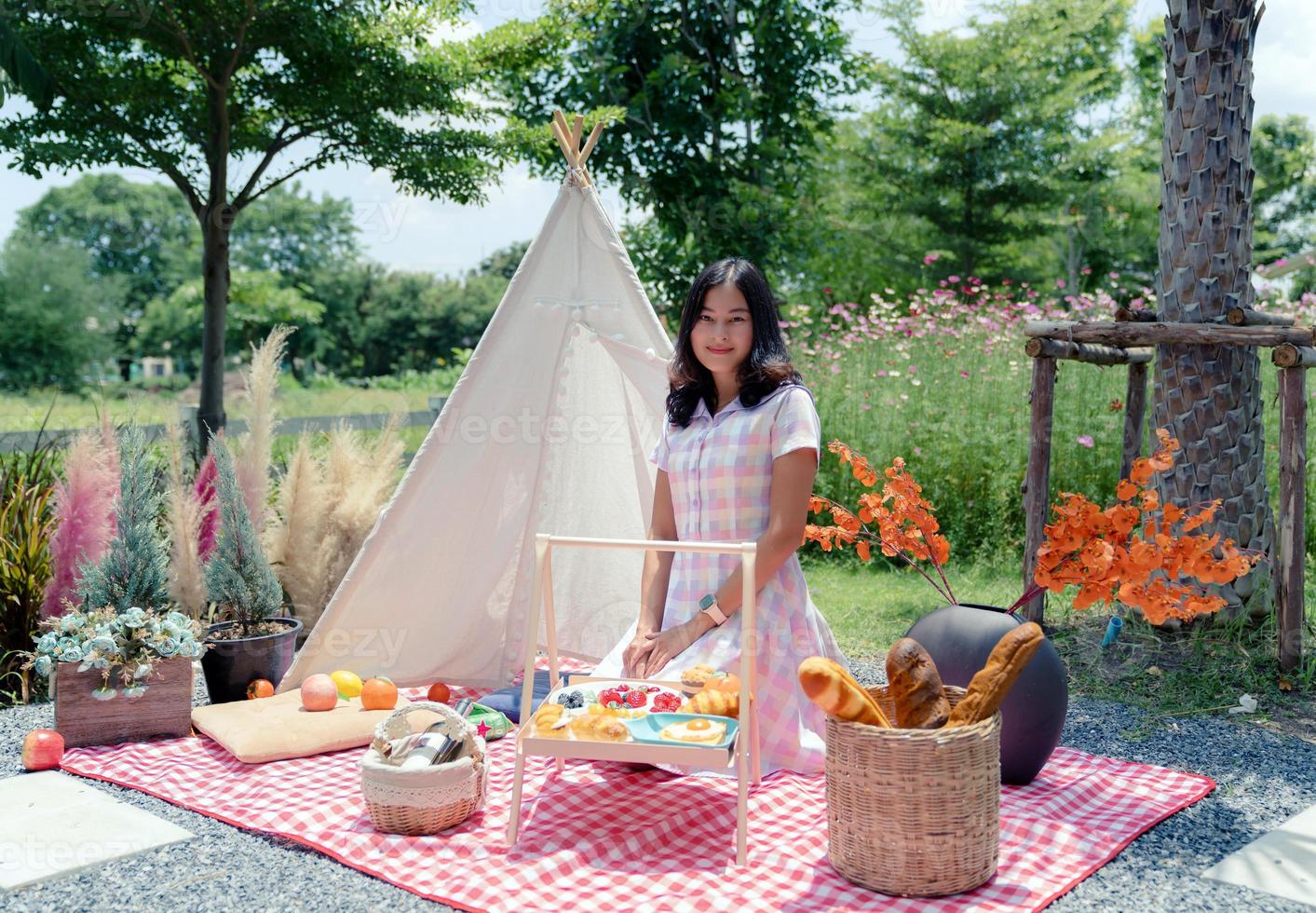 feliz mujer asiática vestida de forma informal sentada en una alfombra de tela con un juego de picnic decorado con una carpa blanca en el jardín foto