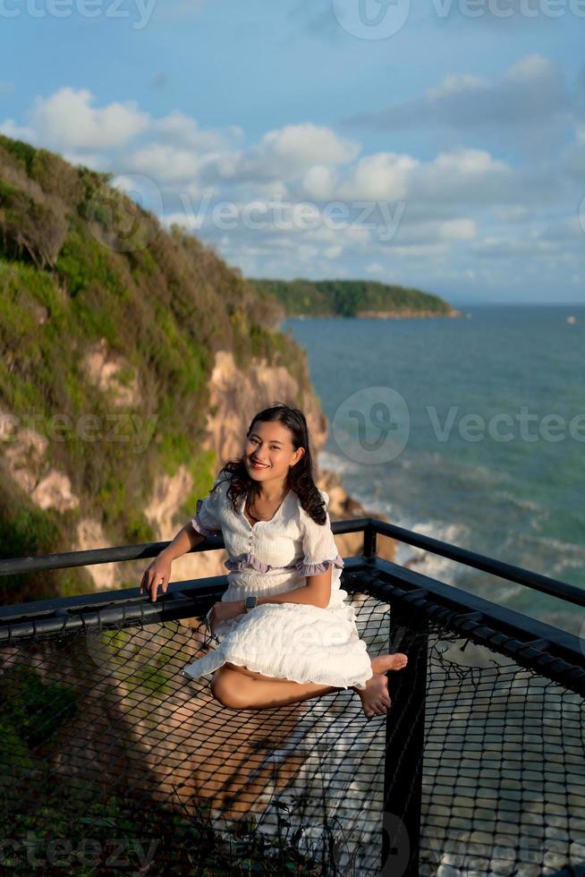 Asian woman tourist sit at net terrace on top hill with view above cliffs and sea in evening with sunlight, sky view photo