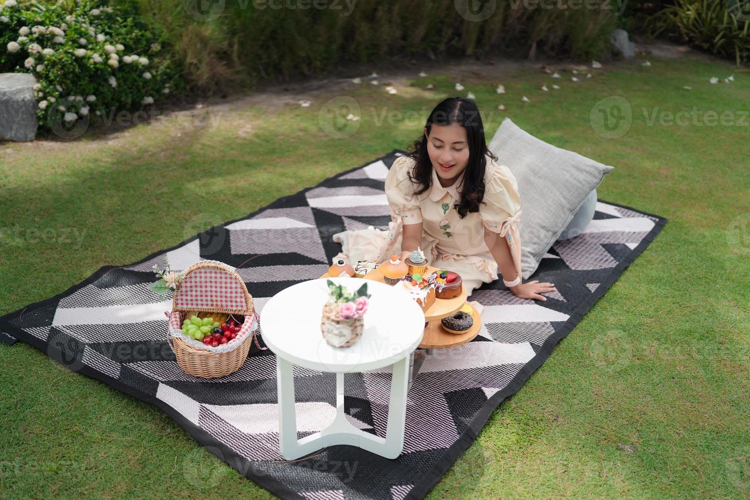 happy Asian woman excited with dessert during picnic in fresh green garden, picnic set with snack and pillow photo