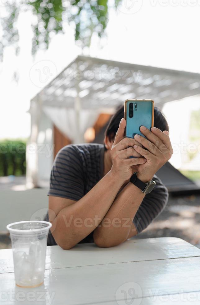 hombre en camiseta causal sosteniendo el teléfono móvil hasta tomar una foto mientras se sienta en una mesa de madera blanca después de tomar café en el jardín de camping, estilo de vida relajado