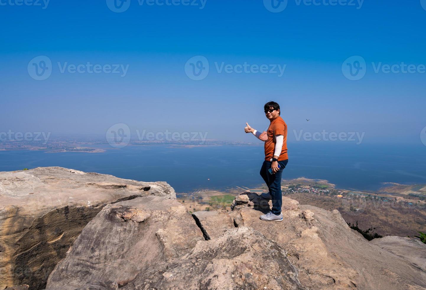 happy Asian man standing with happy smile and thumb up on top viewpoint on rock mountain peak with misty lake dam view and blue sky photo