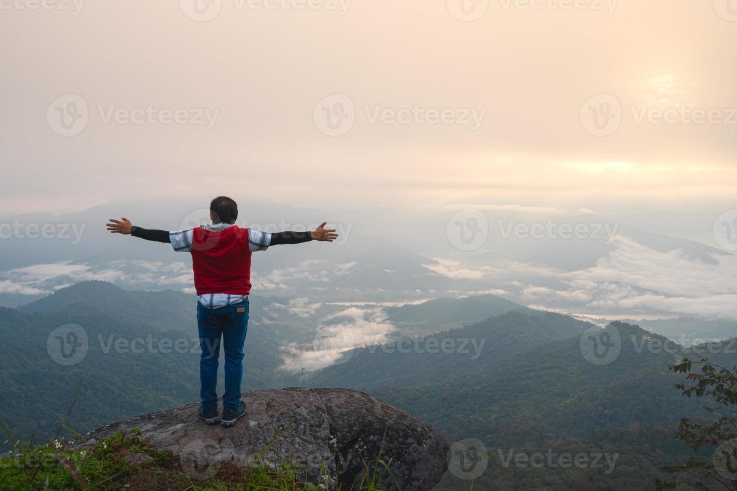 hombre con los brazos libres abiertos de pie en la cornisa rocosa del pico de la montaña en el punto de vista pha tang 104 con vista al río y niebla marina y amanecer en el cielo de niebla pesada. lugar de viaje en chaing rai, tailandia foto