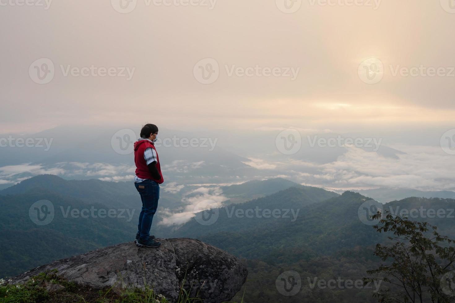 Asian man with protective face mask standing on rock ledge of mountain peak at Pha Tang view point 104 with river view and sea mist and sun rise in heavy fog sky. travel in Chaing Rai, Thailand photo