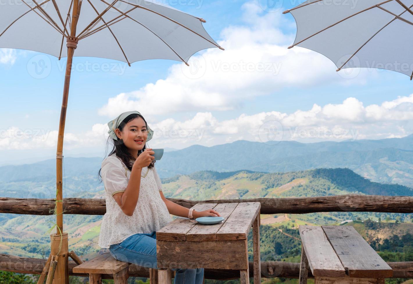 Asian woman relax sit and holding coffee cup at wooden table with white umbrella on terrace with mountain view and cloudy blue sky at Doi Chang, travel attraction in Chiang Rai, Thailand photo